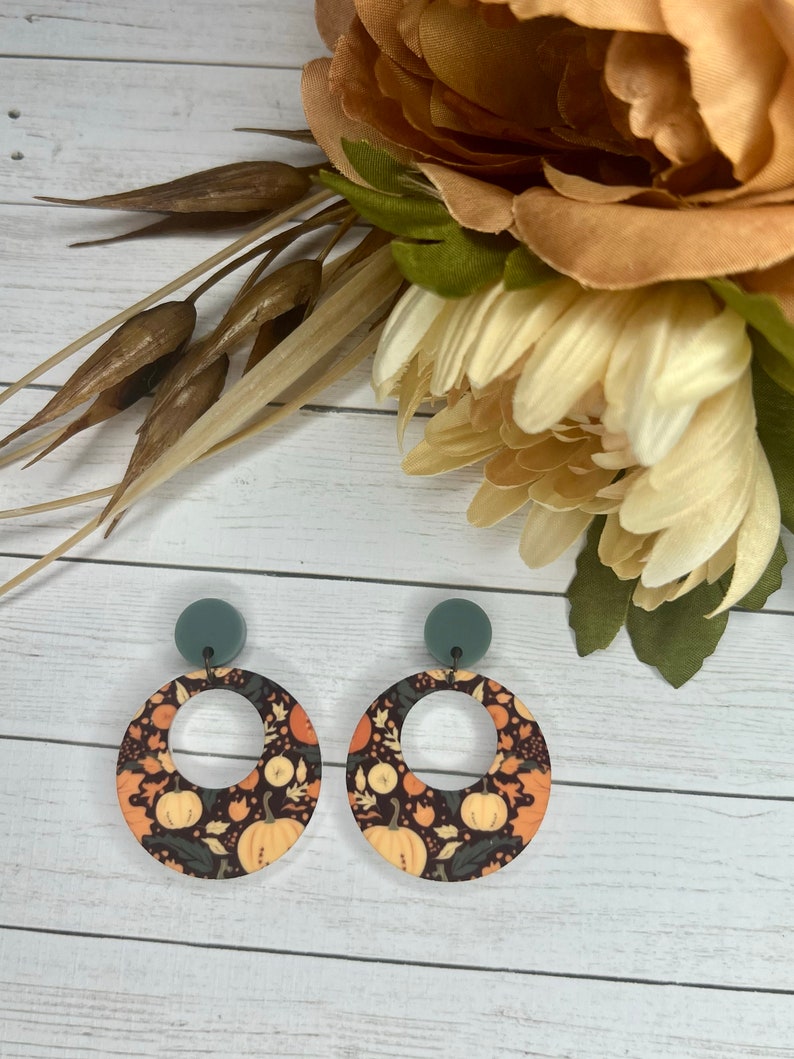 May include: A pair of round, dangle earrings with a black background and a pattern of orange pumpkins, leaves, and flowers. The earrings have a gray top and a silver ring in the center.