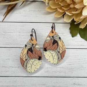 May include: A pair of teardrop-shaped earrings with a fall-themed design. The earrings feature a white background with brown, orange, and yellow leaves and pumpkins.