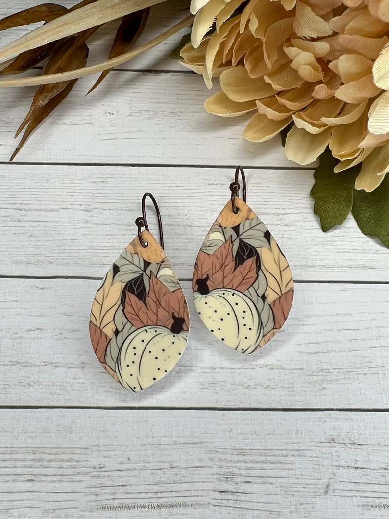 May include: A pair of teardrop-shaped earrings with a fall-themed design. The earrings feature a white background with brown, orange, and yellow leaves and pumpkins.