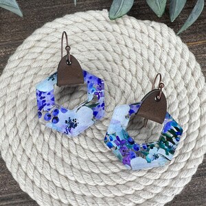 May include: A pair of hexagonal earrings with a floral pattern in shades of purple, blue, and white. The earrings have a brown leather accent and copper wire hooks.