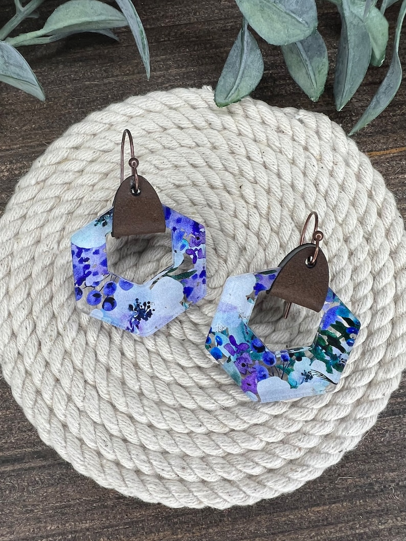 May include: A pair of hexagonal earrings with a floral pattern in shades of purple, blue, and white. The earrings have a brown leather accent and copper wire hooks.