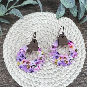 May include: A pair of purple and pink floral earrings with brown leather accents. The earrings are round and have a hole in the center.