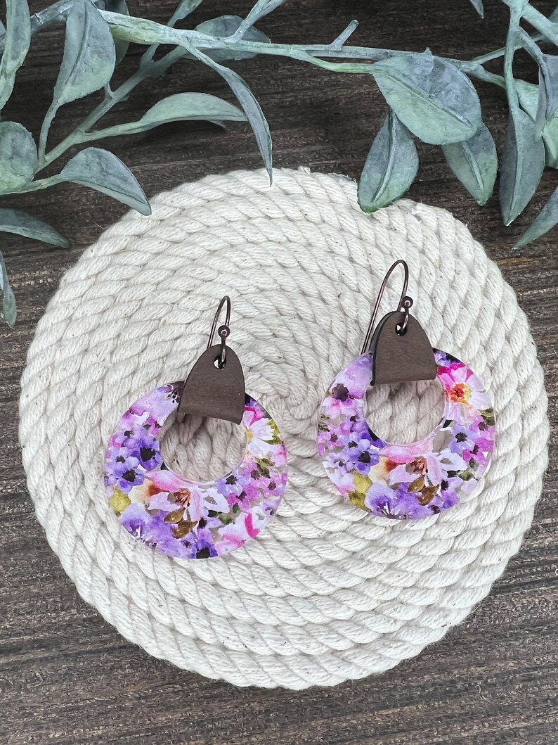 May include: A pair of purple and pink floral earrings with brown leather accents. The earrings are round and have a hole in the center.