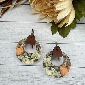 May include: A pair of white and brown circular earrings with a floral and pumpkin design. The earrings have a brown leather accent.