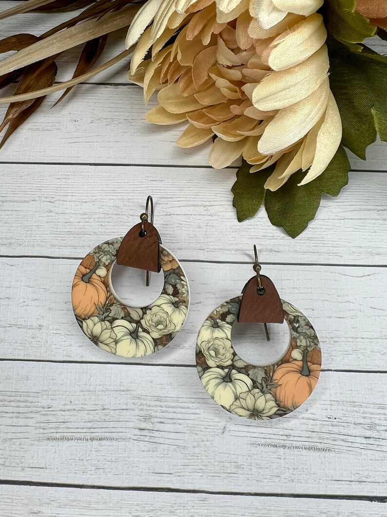 May include: A pair of white and brown circular earrings with a floral and pumpkin design. The earrings have a brown leather accent.