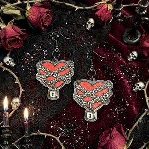 May include: A pair of heart-shaped earrings with a red center, wrapped in a silver chain and secured with a padlock. The earrings hang from silver hooks. The background includes skulls, roses, and candles, creating a gothic aesthetic.