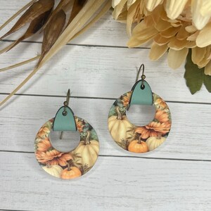 May include: A pair of round, pumpkin-themed earrings with a teal leather accent. The earrings feature a floral design with pumpkins and sunflowers.