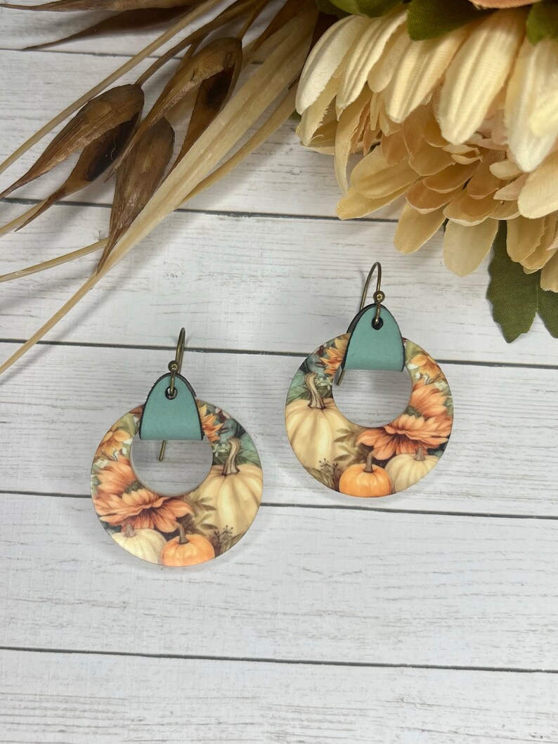 May include: A pair of round, pumpkin-themed earrings with a teal leather accent. The earrings feature a floral design with pumpkins and sunflowers.