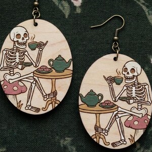 May include: Wooden oval earrings featuring a skeleton enjoying tea at a table with a teapot and cookies. The skeleton is seated on a mushroom. The earrings have bronze-colored ear wires.