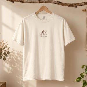 以下が含まれることがあります： オフホワイトの半袖Tシャツで、胸の中央に小さなグラフィックが施されています。グラフィックには、花が咲いた枝にとまる茶色と白の鳥が描かれており、その下に繊細なダークブラウンの筆記体で「Little Things Matter」という文字がプリントされています。