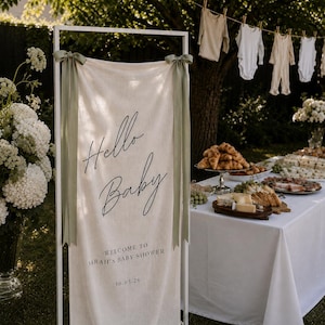 May include: A white rectangular metal frame holding a cream-coloured fabric banner with sage green ribbon ties. The banner features black text reading Hello Baby, Welcome to Sarah's Baby Shower, and the date 30.05.26 in a clean, modern font style.