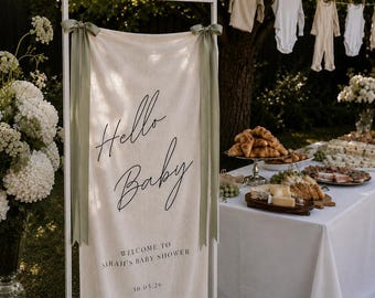May include: A white rectangular metal frame holding a cream-coloured fabric banner with sage green ribbon ties. The banner features black text reading Hello Baby, Welcome to Sarah's Baby Shower, and the date 30.05.26 in a clean, modern font style.