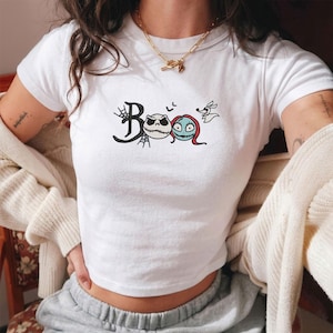 May include: A white short-sleeved t-shirt featuring a graphic print that spells the word Boo with a spiderweb on the B, followed by the faces of Jack Skellington and Sally from The Nightmare Before Christmas, and a small ghost dog character.