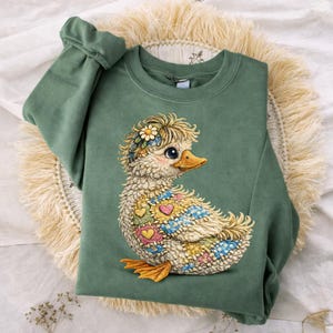 Puede incluir: Una sudadera verde con un diseño de pato bordado. El pato tiene una diadema floral y patrones de corazones y flores coloridos. La sudadera está doblada sobre una estera tejida.