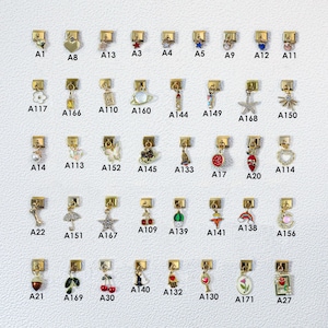 May include: A selection of gold-coloured charms featuring diverse designs such as hearts, stars, and floral motifs. Each charm has a small rectangular clasp for attachment. The charms are arranged in neat rows and columns.