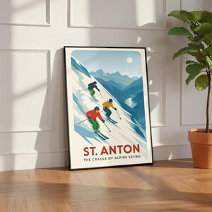 May include: A framed poster featuring a vintage-style illustration of three skiers descending a snowy mountain. The poster has a blue, white, and red color scheme, with the text "ST. ANTON" and "THE CRADLE OF ALPINE SKIING".