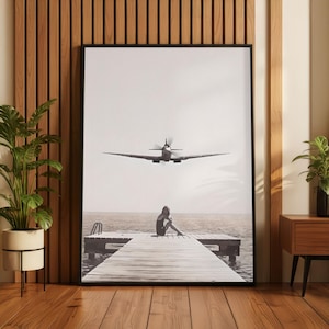 May include: Black and white framed print featuring a vintage airplane flying over a person sitting on a wooden pier. The image has a serene, minimalist aesthetic, suitable for home decor.