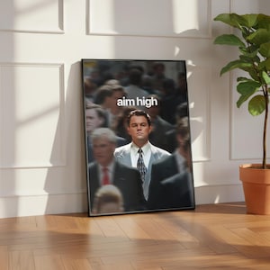 May include: A framed poster with the text "aim high" at the top, featuring a man in a suit. The background is blurred, with other people. The poster is propped against a white wall, with a wooden floor visible.