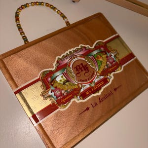 May include: A rectangular wooden cigar box featuring a decorative label with the text My Father Cigars, Garcia & Garcia, and La Lealtad. The box has a handle made of multi-colored beads attached to the top edge, showcasing a natural wood grain finish.
