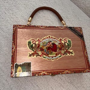 May include: A rectangular handbag crafted from a wooden cigar box with a light brown wood grain finish. It features a braided dark brown handle, a central ornate label depicting figures, and the text Flor de las Antillas and My Father Cigars.