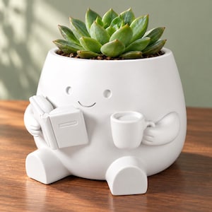 Può includere: Un vaso in ceramica bianca a forma di personaggio seduto che tiene un libro aperto in una mano e una tazza nell'altra. Il vaso presenta una finitura opaca liscia e contiene una pianta succulenta verde con foglie appuntite.