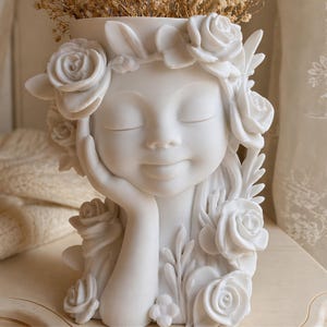 May include: A white ceramic planter shaped like a serene face, adorned with rose details and a floral crown. The figure's hand gently supports the chin. The planter is filled with dried flowers, creating a decorative piece for home decor.