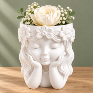 Könnte beinhalten: Ein weißer Keramikübertopf in Form eines Kopfes mit geschlossenen Augen und Händen unter dem Kinn. Die Figur trägt einen modellierten Blumenkranz. Der Übertopf ist mit einer großen weißen Blüte und kleinen weißen Schleierkrautblüten gefüllt.