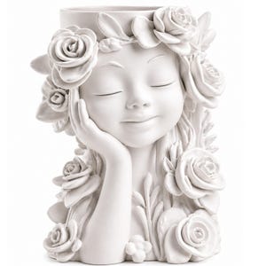 May include: A white ceramic planter in the shape of a girl's face, decorated with roses. The girl has her eyes closed and her chin resting on her hand. Roses are sculpted around her head and hair, forming a floral crown.
