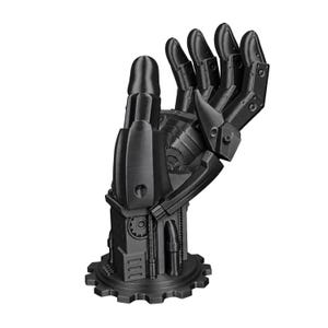 May include: A matte black robotic hand sculpture featuring articulated finger segments and mechanical detailing. The hand is mounted on a circular gear-shaped base, showcasing intricate joints, exposed gears, and cylindrical components throughout the structure.