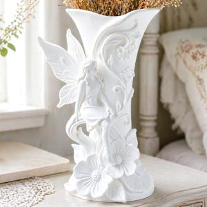 Può includere: Un vaso in ceramica bianca con un design a forma di fata. Il vaso presenta una fata seduta con grandi ali, circondata da dettagli floreali e di foglie. Il vaso è pieno di fiori secchi ed è alto circa 30 cm.