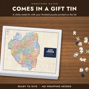 Op de afbeelding: Een witte metalen geschenkblik met een kaart van Ann Arbor, Michigan, op het deksel gedrukt. De tekst luidt HOMETOWN MAVEN, COMES IN A GIFT TIN, A white metal tin with your finished puzzle printed on the lid, ANN ARBOR MICHIGAN, READY TO GIVE, NO WRAPPING NEEDED.