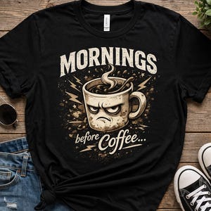 May include: Black t-shirt featuring a grumpy coffee cup graphic with the text "Mornings before Coffee...". The design includes lightning bolts and a coffee steam effect. The shirt is displayed on a wooden surface with sunglasses, jeans, and sneakers.