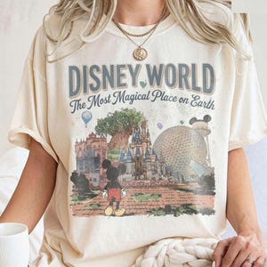 Puede incluir: Camiseta color crema con el texto "DISNEY WORLD The Most Magical Place on Earth" en azul y blanco. El gr&aacute;fico incluye ilustraciones de lugares emblem&aacute;ticos de Disney y Mickey Mouse. La camiseta tiene mangas cortas.