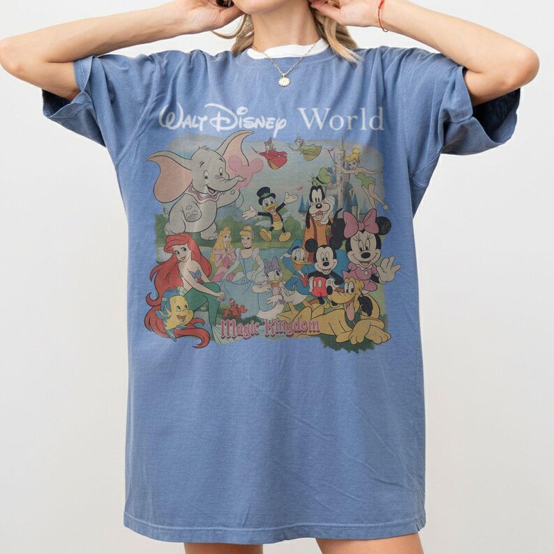 Puede incluir: Una camiseta azul de manga corta con un estampado gr&aacute;fico de varios personajes de Disney, incluidos Dumbo, Ariel, Mickey Mouse y Minnie Mouse. El texto "Walt Disney World" y "Magic Kingdom" est&aacute; impreso encima y debajo de la ilustraci&oacute;n.