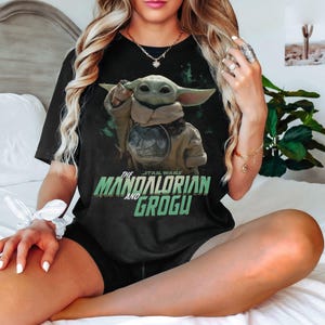 May include: Black t-shirt featuring Grogu from The Mandalorian. The graphic shows Grogu with a tan cloak and the text "The Star Wars The Mandalorian and Grogu" in green and white. The shirt is worn by a person.