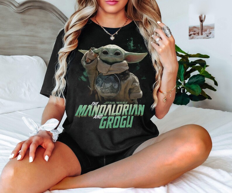 May include: Black t-shirt featuring Grogu from The Mandalorian. The graphic shows Grogu with a tan cloak and the text "The Star Wars The Mandalorian and Grogu" in green and white. The shirt is worn by a person.