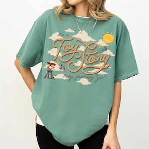 Op de afbeelding: Een saliegroen T-shirt met korte mouwen en een afbeelding van Woody en Jessie uit Toy Story. Het ontwerp bevat tekst in touwstijl met "it's a Toy Story" en "Baby, just say 'yes'" tegen een achtergrond van witte wolken en een gele zon.