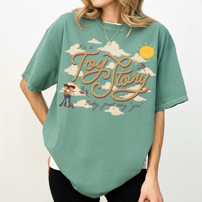 Op de afbeelding: Een saliegroen T-shirt met korte mouwen en een afbeelding van Woody en Jessie uit Toy Story. Het ontwerp bevat tekst in touwstijl met "it's a Toy Story" en "Baby, just say 'yes'" tegen een achtergrond van witte wolken en een gele zon.