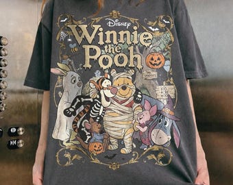 May include: A charcoal gray short-sleeve t-shirt featuring a Disney Winnie the Pooh Halloween graphic. The print shows Pooh as a mummy, Tigger in a skeleton costume, Piglet as a vampire, and Eeyore in a witch hat, surrounded by ornate gold filigree.