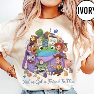 May include: An ivory t-shirt featuring a graphic print of various animated characters from Toy Story emerging from a purple backpack. The text "You're Got a Friend In Me" is printed in blue below the characters. A circular "IVORY" label is in the corner.