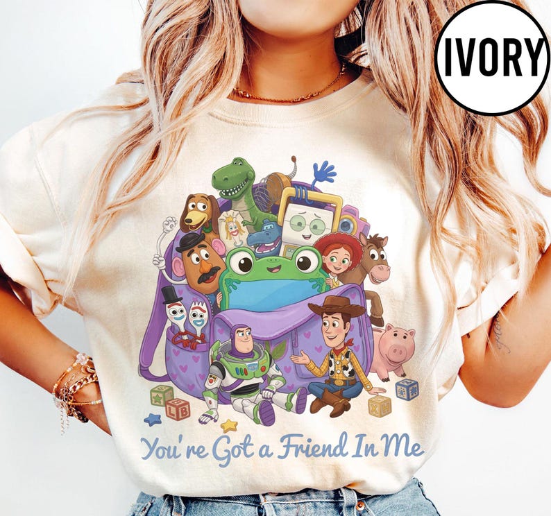 May include: An ivory t-shirt featuring a graphic print of various animated characters from Toy Story emerging from a purple backpack. The text "You're Got a Friend In Me" is printed in blue below the characters. A circular "IVORY" label is in the corner.