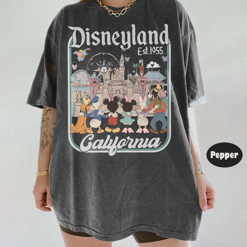 Puede incluir: Camiseta gris oscuro con un gr&aacute;fico de Disneyland. El dise&ntilde;o incluye el logotipo de Disneyland, el texto "Est. 1955" y una colorida ilustraci&oacute;n de personajes de Disney y el castillo. Tambi&eacute;n est&aacute; presente la palabra "California".