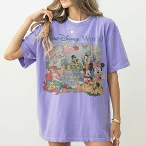 Puede incluir: Una camiseta de manga corta de color morado claro con un estampado gr&aacute;fico de varios personajes de Disney, incluidos Dumbo, Ariel, Cenicienta, Mickey Mouse, Minnie Mouse y Pluto, con el texto Walt Disney World y Magic Kingdom en el frente.