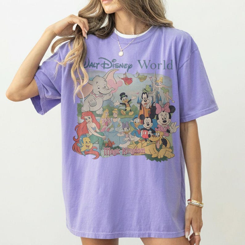 Puede incluir: Una camiseta de manga corta de color morado claro con un estampado gr&aacute;fico de varios personajes de Disney, incluidos Dumbo, Ariel, Cenicienta, Mickey Mouse, Minnie Mouse y Pluto, con el texto Walt Disney World y Magic Kingdom en el frente.