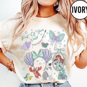 May include: An ivory t-shirt featuring a graphic print with the text Part Of Your World, a mermaid character in a mirror frame, a crab, a fish, a dog, a butterfly, bubbles, and a headband with seashell ears and a bow in pastel colors.