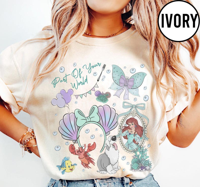 May include: An ivory t-shirt featuring a graphic print with the text Part Of Your World, a mermaid character in a mirror frame, a crab, a fish, a dog, a butterfly, bubbles, and a headband with seashell ears and a bow in pastel colors.