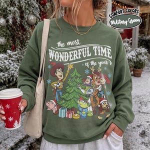 May include: A military green crewneck sweatshirt featuring a graphic print of Toy Story characters decorating a Christmas tree. The text reads, "the most WONDERFUL TIME of the year." The sweatshirt includes a beige shoulder bag strap.