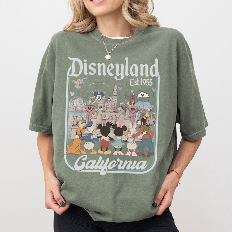 Puede incluir: Camiseta verde oliva con un gr&aacute;fico de Disneyland. El gr&aacute;fico incluye el logotipo de Disneyland, el texto "Est.1955" e ilustraciones de personajes de Disney y el castillo. Tambi&eacute;n se incluye la palabra "California".