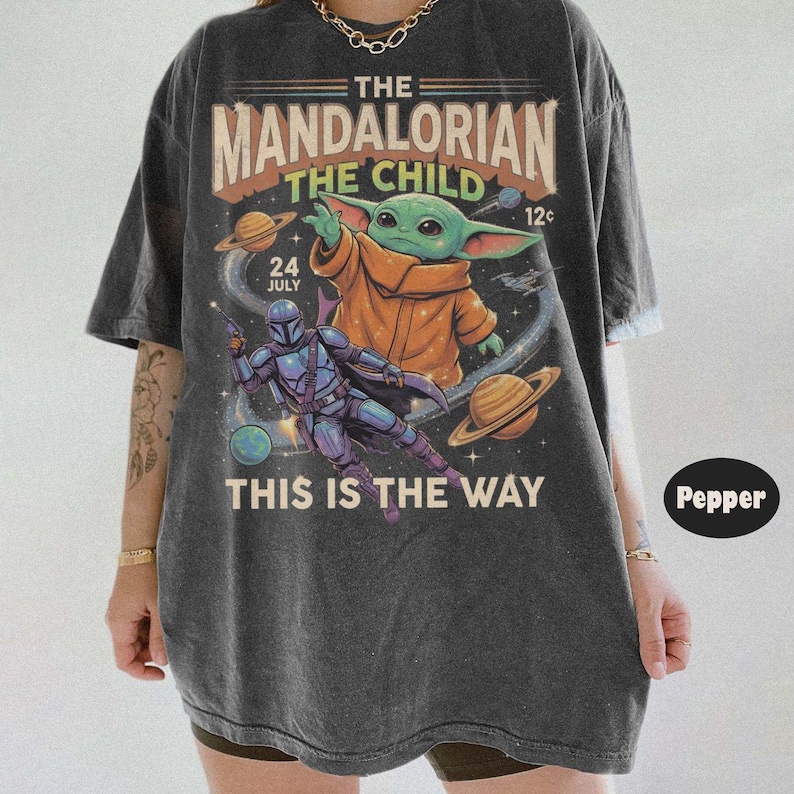 Op de afbeelding: Een antracietgrijs t-shirt met korte mouwen en een grafische print van The Mandalorian en The Child in de ruimte. De tekst luidt THE MANDALORIAN THE CHILD 24 JULY 12c THIS IS THE WAY. Het ontwerp bevat planeten en sterren in vintage stijl.