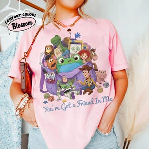 May include: A light pink t-shirt featuring a graphic print of various animated toy characters emerging from a purple backpack. The text "You're Got a Friend In Me" is printed in blue below the graphic. The shirt has short sleeves and a crew neckline.